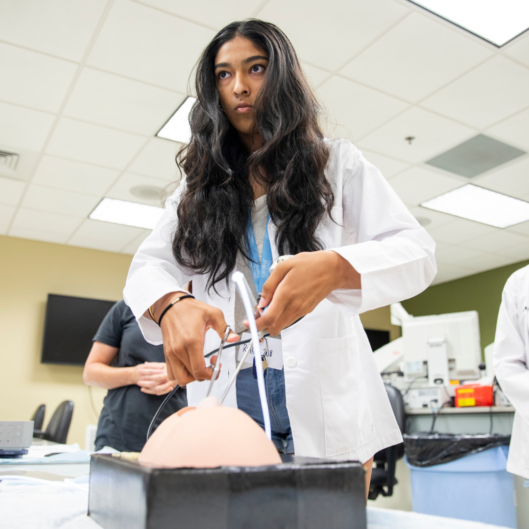A person in a lab coat operates medical instruments on a training model, in a brightly lit clinical lab with people and equipment in the background.