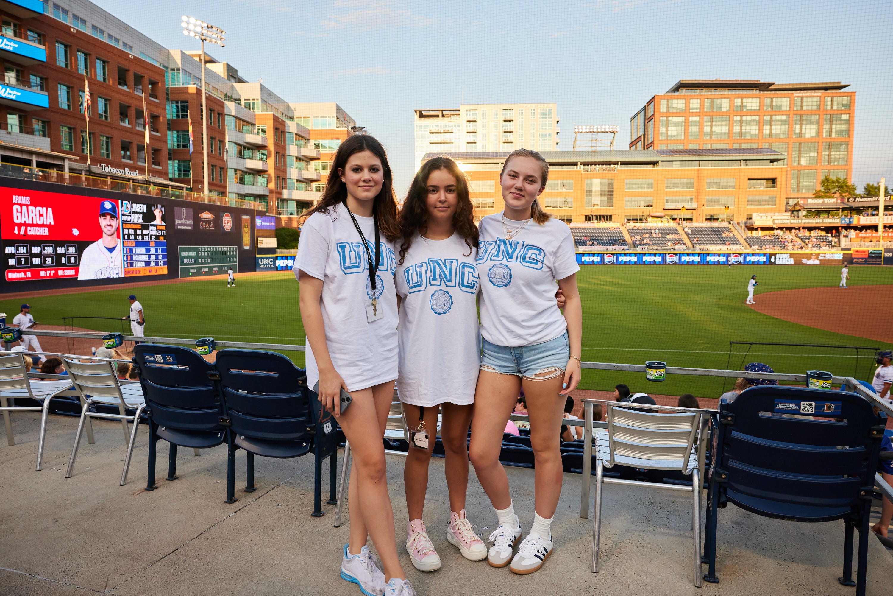 Three students wearing "UNC" shirts stand in front of a baseball field, with a scoreboard displaying "Aramis Garcia, 4, Catcher" and buildings in the background during daylight.