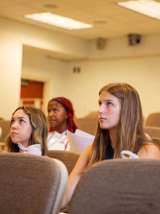 Three students sit attentively in a lecture hall, focusing forward. They hold papers, surrounded by rows of brown seats, with a lit ceiling visible above.
