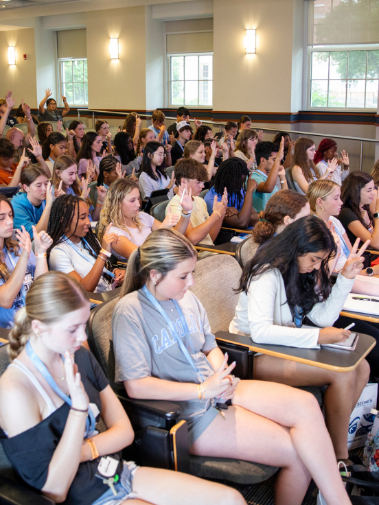 A diverse group of students interested in medicine sit attentively in a classroom-like setting, many raising their hands, engaging in discussion. The room is bright, with large windows and overhead lighting.