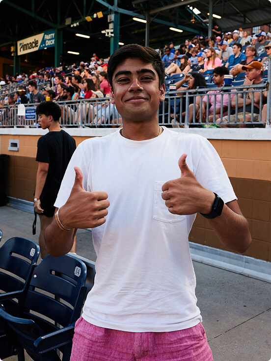 A future medical student gives two thumbs up, smiling in a white shirt and pink shorts, standing in front of stadium seating filled with spectators. The text "Pinnacle Party Deck" is visible.