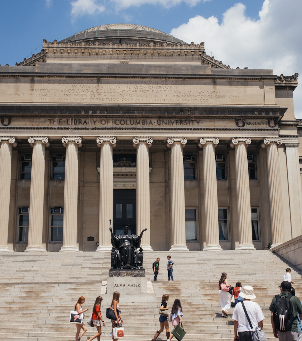 A statue labeled "Alma Mater" sits before Columbia University's library, featuring grand columns and a dome. People walk on the steps under a blue sky, engaging in conversation.