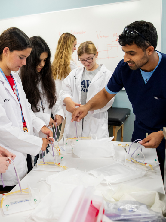 A group of people in lab coats participate in a hands-on activity at a table in a classroom. A person in scrubs guides them; a whiteboard shows diagrams behind them.