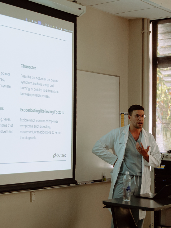 A doctor talks beside a projected presentation slide, detailing medical symptoms and factors, in a classroom with a window. Items on the table include a water bottle and sanitizer. Text on slide:
 

 - "Character
  Describe the nature of the pain or symptom, such as sharp, dull,
  burning, or colicky, to differentiate between possible causes.
 

 - Exacerbating/Relieving Factors
  Explore what worsens or improves symptoms, such as eating, movement, or medications, to refine the diagnosis.
 

 - Outset"