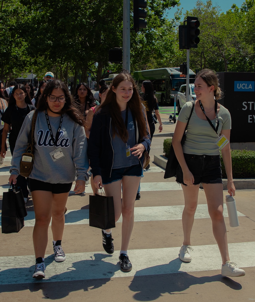 Three young women walk across a crosswalk, each holding bags. Trees and a bus are visible in the background. A sign reads, "UCLA STORE EVENTS."