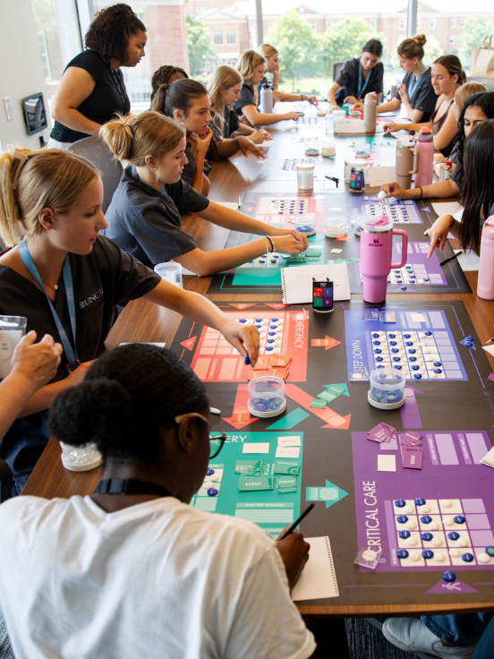 Participants engage in a tabletop exercise at a conference room, using colorful game boards and charts labeled "Emergency," "Surgery," "Vertical Care," and "Step Down," while taking notes and interacting.