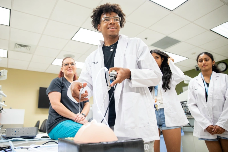 A future medical student in a lab coat manipulates a medical device attached to a silicone model, watched by a seated woman and two standing individuals, in a brightly lit laboratory.