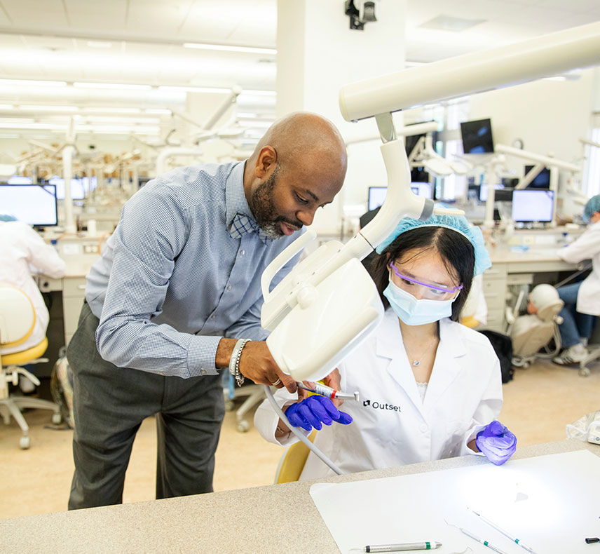 A doctor in a blue shirt assists a student wearing a lab coat and gloves with dental equipment in a classroom setting, surrounded by dental chairs and monitors.
